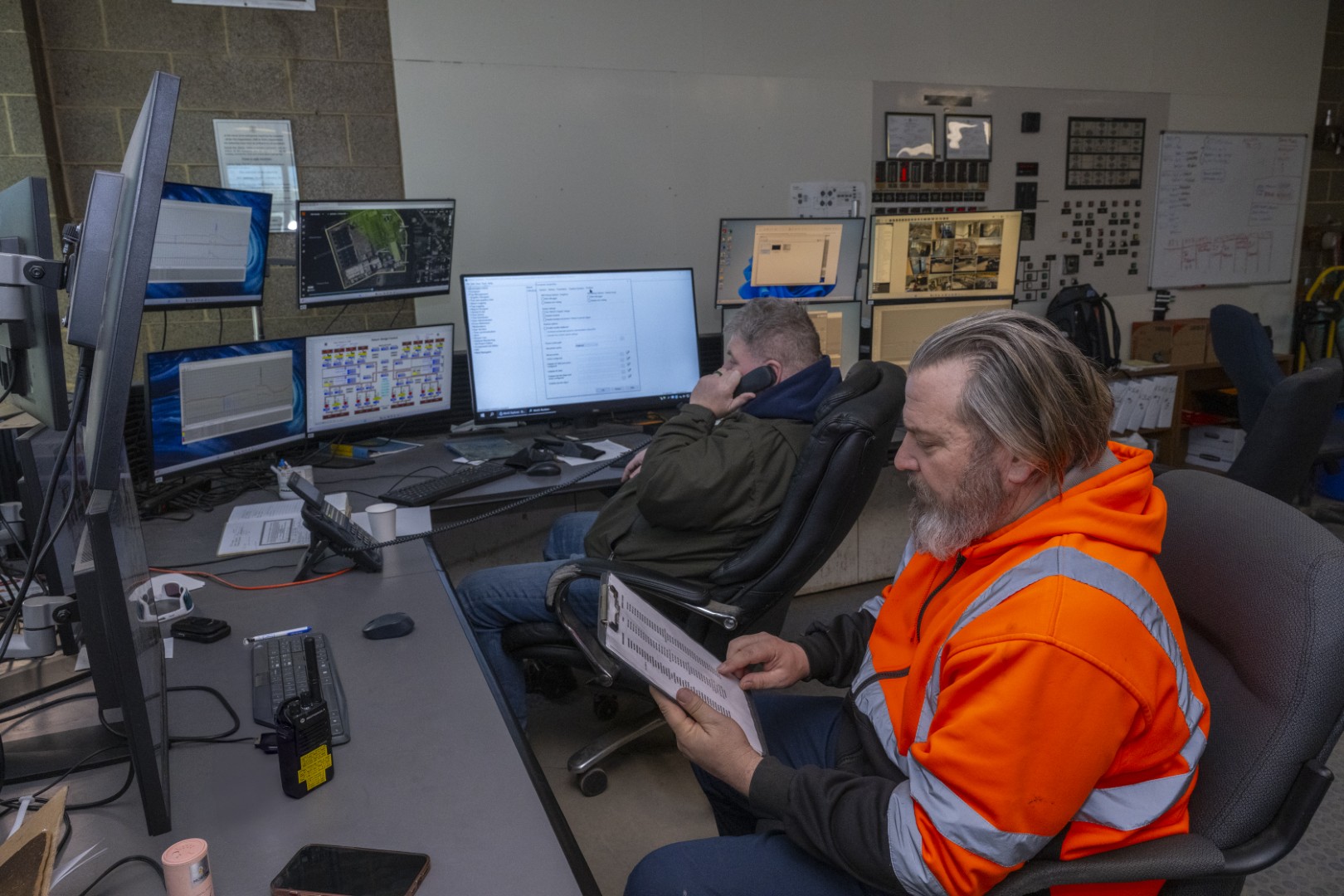 Two workers at a desk with multiple monitors displaying maps, data, and surveillance footage in a control room.