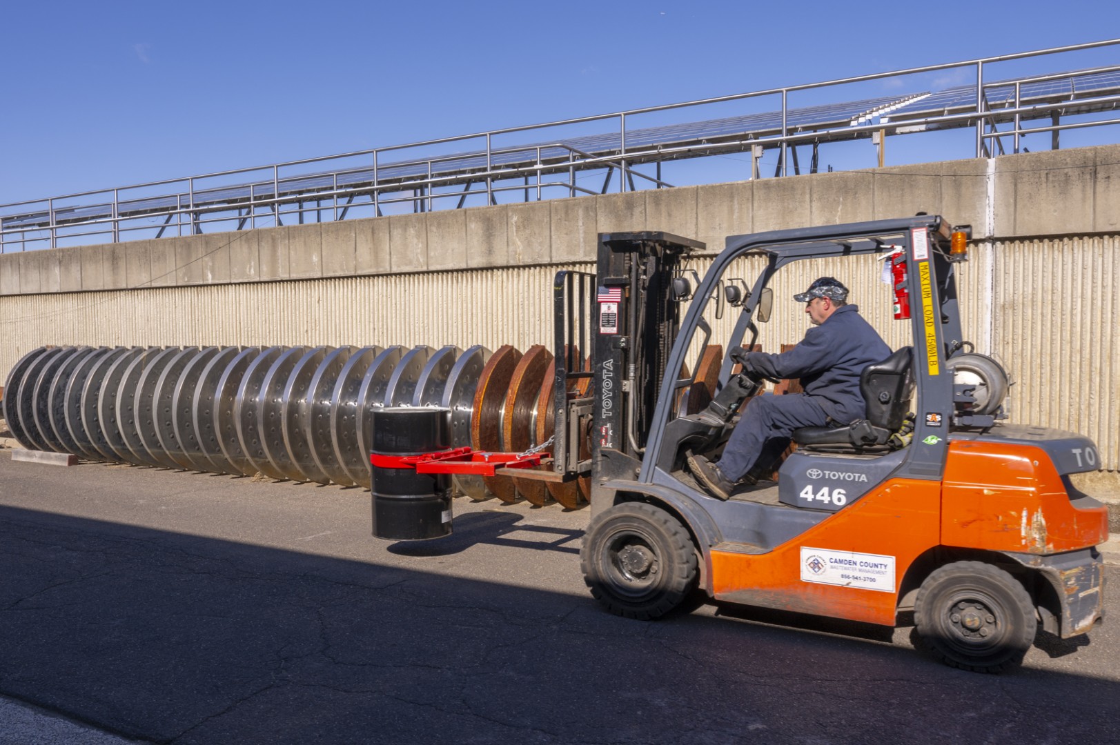 Worker driving a Toyota forklift carrying a black barrel with a red clamp attachment.